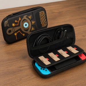 Puede incluir: Estuche de transporte negro con cremallera para consola de juegos, con un diseño marrón y dorado en el exterior. El estuche está abierto, revelando un mando azul y rojo, cables y compartimentos.