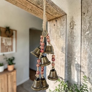 Puede incluir: Un carillón de viento colgante con seis campanas de color bronce suspendidas de una cuerda natural. Las campanas están ensartadas con cuentas rojas y marrones y una moneda decorativa. Para uso en interiores o exteriores.