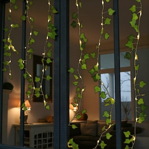 Op de afbeelding: Decoratieve klimop-slingerverlichting met groene bladeren en kleine, warmwitte LED-lampjes. De lampjes zijn verticaal tegen een raam geplaatst, wat een natuurlijke en uitnodigende sfeer creëert. Perfect voor binnen- of buitendecoratie.
