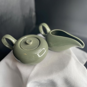 May include: A sage green ceramic teapot with a matching lid and a curved handle, paired with a matching sage green ceramic creamer pitcher featuring a pointed spout and a side handle. Both pieces have a smooth, glossy finish and rounded shapes.