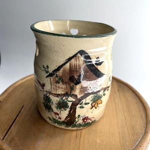 May include: A cylindrical ceramic vessel with a cream-colored speckled glaze, featuring a hand-painted brown house nestled in a tree with green foliage and small red and yellow flowers. The rim has a dark green border and heart-shaped cutouts.