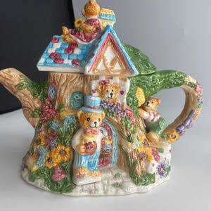 May include: A ceramic teapot shaped like a cottage with a tree trunk base, featuring multiple teddy bears. One bear wears a blue top hat, while others are positioned on the roof and side. The design includes colorful flowers, green foliage, and a blue-checkered roof.