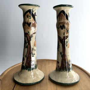May include: A pair of cream-colored ceramic candle holders with a glossy finish. Each holder features a hand-painted design of a brown tree with green leaves and a dark house silhouette at the top. The base and rim are trimmed in dark green.
