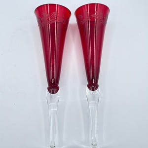 May include: Two tall champagne flutes featuring deep red glass bowls with etched holly leaf and berry patterns near the rims. Each glass has a clear, slender stem and a circular base, providing a contrast to the vibrant red upper section.