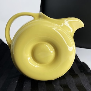 May include: A bright yellow ceramic pitcher with a smooth, glossy finish. The vessel features a circular, donut-shaped body with a central indentation, a curved handle on the left side, and a wide, pointed pouring spout on the right side.