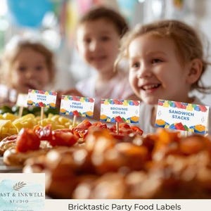 May include: Four rectangular party food labels on wooden picks featuring a colorful plastic building block pattern. The labels display the text VEGGIE STICKS, FRUIT TOWERS, BROWNIE BRICKS, and SANDWICH STACKS in blue font against a white background.