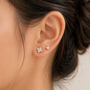 May include: Close-up of a person's ear with two sets of gold stud earrings. One set features a cluster of four small, clear gemstones in a square shape. The other set has a single flower shape with clear gemstones.