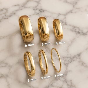 Peut inclure: Six anneaux dorés disposés sur deux rangées, présentant des largeurs de bande variables étiquetées 6 mm, 5 mm, 4 mm, 3 mm, 2 mm et 1 mm. Chaque anneau possède une surface lisse, polie et réfléchissante, montrant une gamme d'épaisseurs.