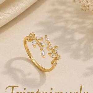 Könnte beinhalten: Ein goldfarbener offener Ring mit einem zarten Zweigdesign, das mit kleinen, klaren, im Marquise- und Rundschliff geschliffenen Steinen besetzt ist. Der Text Triptajewels und der Schriftzug CRAFTED WITH LOVE, MADE TO SHINE stehen unter dem Ring.