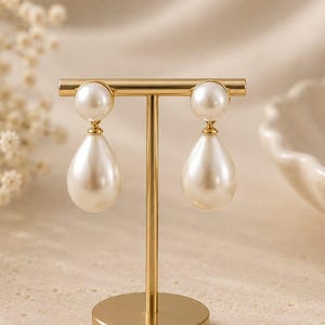 May include: A pair of gold-toned drop earrings featuring a small round pearl stud connected to a larger teardrop-shaped pearl pendant. The earrings are displayed on a gold-colored T-shaped metal stand with a circular base and a smooth finish.