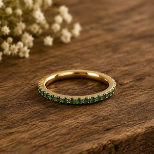May include: A thin gold band ring featuring a continuous row of small, round, vibrant green gemstones set along the entire circumference of the metal band, creating a delicate and uniform circular design with a polished metallic finish.