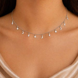 May include: A silver necklace featuring charms shaped like stars, moons, and lightning bolts. The necklace is worn around the neck and hangs against a neutral background. The jewelry is delicate and stylish.
