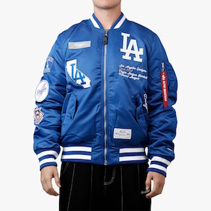 Może przedstawiać: Niebieska satynowa kurtka bomberka z białymi akcentami i wieloma naszywkami. Kurtka ma logo i nazwę drużyny Los Angeles Dodgers. Kurtka ma zapięcie na zamek błyskawiczny oraz prążkowane mankiety i dół z białymi paskami.