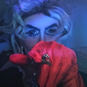 Puede incluir: Una persona con maquillaje dramático y una peluca plateada señala al espectador. Llevan un guante rojo con un anillo plateado. El fondo es azul oscuro con un toque de morado.