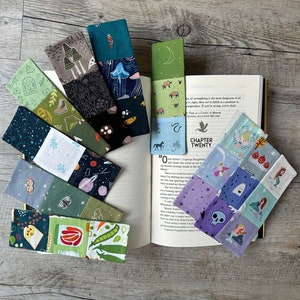 Può includere: Una collezione di 10 segnalibri in tessuto colorati con diversi motivi. I segnalibri sono realizzati in tessuto patchwork con vari disegni, tra cui funghi, stelle e un teschio. I segnalibri sono disposti su una superficie di legno con un libro aperto a una pagina con il titolo "Capitolo Venti".