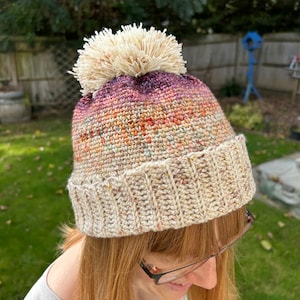 May include: A crocheted beanie with a white pom pom. The beanie is a gradient of colors, starting with a deep purple at the top and fading to a light beige at the bottom.