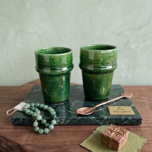 Könnte beinhalten: Zwei grüne Keramikbecher auf einem grünen Marmortablett, zusammen mit einem Kupferlöffel und einem Jade-Perlenarmband. Ein kleiner Holzstempel und eine Messingplakette sind ebenfalls vorhanden. Die Becher haben eine strukturierte, glänzende Oberfläche.