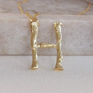 May include: Gold-colored initial "H" pendant necklace. The pendant has a textured, organic design, resembling tree bark. The necklace chain is delicate and gold-toned. The pendant is displayed on a light-colored surface.