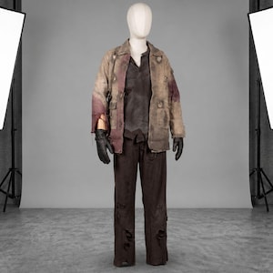 May include: A costume on a mannequin, featuring a distressed tan jacket with burgundy accents, a dark brown shirt, and matching pants. The outfit includes black gloves and is displayed in a studio setting with lighting equipment.