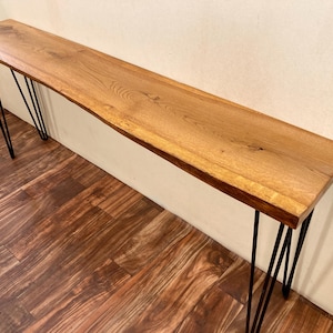 May include: A long, rectangular wooden console table with a natural wood finish. The table is supported by black metal hairpin legs, creating a mid-century modern aesthetic. The wood grain is visible, adding to the table's rustic charm.