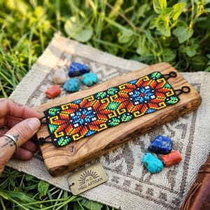 May include: A colorful beaded bracelet with a geometric floral pattern in red, orange, blue, green, and yellow. The bracelet is displayed on a wooden board with a small tag that reads "H&G Artisan".