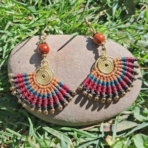 Peut inclure: Une paire de boucles d'oreilles en forme d'éventail avec du cordon macramé dans des tons bordeaux, bleu sarcelle et beige. Chaque boucle comprend une spirale en laiton, une perle rouge-brun et des petites perles dorées sur le bord inférieur.