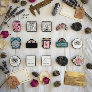 Könnte beinhalten: Eine Sammlung von Emaille-Anstecknadeln mit verschiedenen Motiven, darunter eine rosa Limonadendose, Gehirnillustrationen, chemische Strukturen für Serotonin und Dopamin sowie Text-Pins wie 'Weird Girls Club' und 'Dopamine ADHD Switch'.