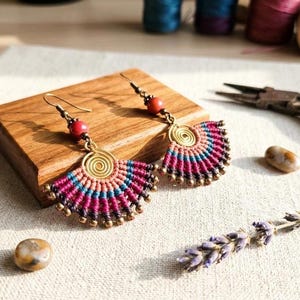 Peut inclure: Une paire de boucles d'oreilles en macramé en forme d'éventail avec une spirale centrale en laiton, tissées avec du cordon rose, bleu et violet foncé. Chaque boucle est ornée d'une perle rouge et de petites perles en laiton sur le bord inférieur.