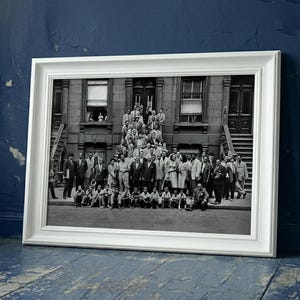 May include: A framed black and white photograph featuring a large group of people posing in front of a building. The photograph is in a white frame and is leaning against a blue wall.