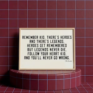 May include: A framed print with a light wood frame displays black text on a white background. The text reads, "REMEMBER KID. THERE'S HEROES AND THERE'S LEGENDS. HEROES GET REMEMBERED BUT LEGENDS NEVER DIE. FOLLOW YOUR HEART KID. AND YOU'LL NEVER GO WRONG." The print is on a red tiled surface.