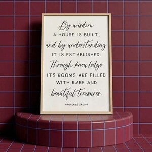 Puede incluir: Una impresión enmarcada con un marco de madera clara. La impresión presenta una elegante escritura negra sobre un fondo blanquecino. El texto dice: "Con la sabiduría se edifica la casa, y con la inteligencia se afirma." Proverbios 24:3-4.