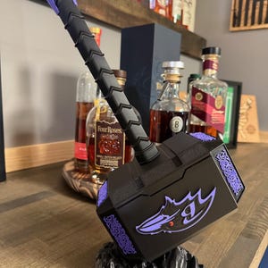 May include: A black and purple decorative hammer with a stylized dragon emblem. The hammer features a textured handle and intricate purple detailing. It rests on a black, rocky base, and is displayed on a wooden surface.