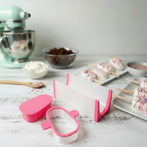 May include: A pink and white popsicle mold with a lid and a popsicle-shaped cutter. The image also shows a white and pink popsicle holder, a mint green stand mixer, and decorated popsicles on a white plate.