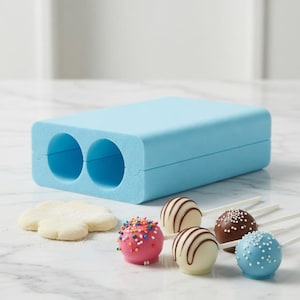 以下が含まれることがあります： 2つの円形キャビティを備えた、水色のケーキポップ型。 ピンクのスプリンクル、チョコレートストライプの白、白いスプリンクルのチョコレートなど、いくつかの装飾されたケーキポップが表示されています。 花の形のクッキーカッターも存在します。