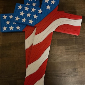 Op de afbeelding: Een houten kruis met het ontwerp van de Amerikaanse vlag. Het kruis heeft een blauw gedeelte met witte sterren en rood-witte strepen. Het kruis is gemaakt van hout en is een patriottische decoratie.