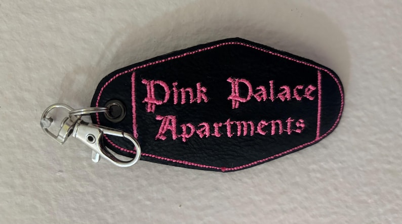 Może przedstawiać: Czarny brelok z wyhaftowanymi r&oacute;żowymi słowami "Pink Palace Apartments". Brelok ma srebrne zapięcie i r&oacute;żową obw&oacute;dkę. Brelok jest brelokiem w stylu hotelowym.