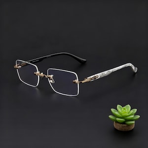 May include: Rimless eyeglasses with square lenses, gold-colored metal accents, and black temples. The arms feature a white and gray marble pattern. The glasses are displayed on a dark background.