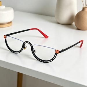 May include: Black and red half-rim eyeglasses with a unique design. The frames feature a black metal structure with a distinctive curved shape around the lenses. The temples and tips are red, and the frames have orange accents.