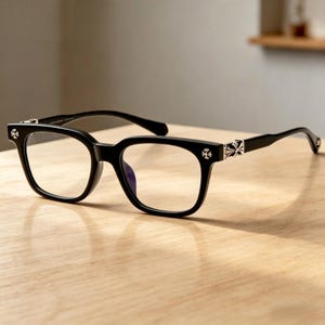 May include: Black square-framed spectacles with clear lenses. The frames have decorative silver cross accents on the temples and hinges. The glasses are set on a light-coloured wooden surface.