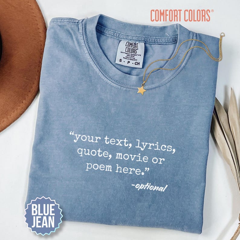 以下が含まれることがあります： クルーネックの半袖デニムブルーTシャツ。シャツには「your text, lyrics, quote, movie or poem here.」および「-optional」という白いテキストがプリントされています。襟元には星のペンダントが付いたゴールドのネックレスが置かれています。