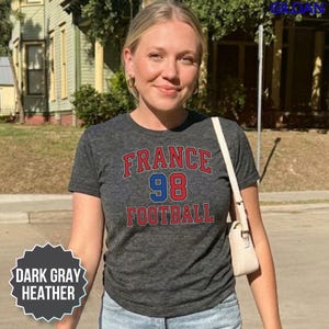 A smiling woman stands on a sunny street wearing a dark gray heather France soccer shirt with a classic 90s retro print. This comfortable Y2K style tee pays a subtle tribute to the historic 1998 world championship victory, making it perfect for today's 2026 tournament buzz. Whether you are meeting your group at a sports pub or cheering from the arena bleachers, it delivers a stylish fan look. Order now to get fast domestic delivery across the US.