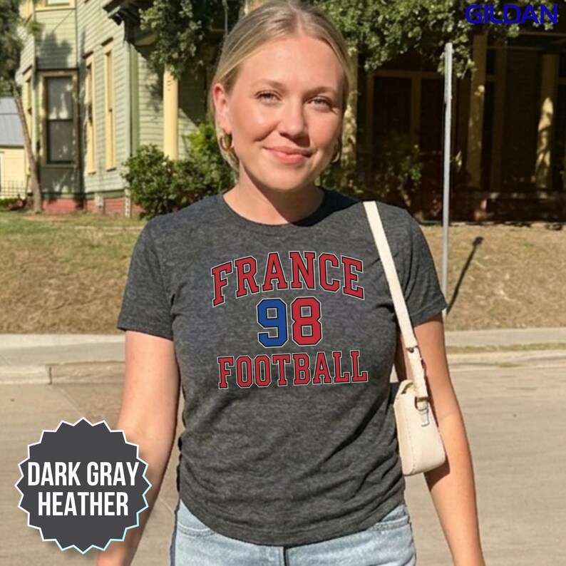 A smiling woman stands on a sunny street wearing a dark gray heather France soccer shirt with a classic 90s retro print. This comfortable Y2K style tee pays a subtle tribute to the historic 1998 world championship victory, making it perfect for today's 2026 tournament buzz. Whether you are meeting your group at a sports pub or cheering from the arena bleachers, it delivers a stylish fan look. Order now to get fast domestic delivery across the US.