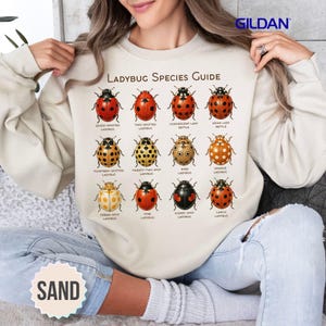 Può includere: Una felpa girocollo color sabbia con una stampa grafica intitolata Ladybug Species Guide. Il design mostra dodici illustrazioni di coccinelle disposte in una griglia, ognuna etichettata con il proprio nome in testo nero.