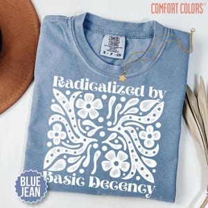 Op de afbeelding: Een blauw spijkerkleurig t-shirt met een witte grafische print met de tekst "Radicalized by Basic Decency" omringd door gestileerde bloemen- en bladpatronen. Het shirt heeft een ronde hals en een Comfort Colors merklabel in de kraag.