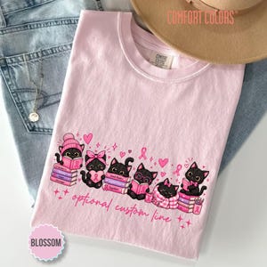 Puede incluir: Una camiseta rosa claro con un gráfico de seis gatos negros con accesorios rosas, libros y lazos de concienciación sobre el cáncer de mama. El texto "optional custom line" está impreso debajo de los gatos. Tiene una etiqueta Comfort Colors.