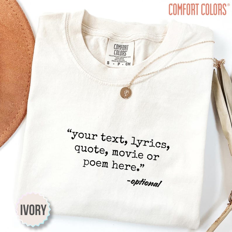 以下が含まれることがあります： クルーネックのアイボリー色の半袖Tシャツ。シャツには黒い文字で「your text, lyrics, quote, movie or poem here.」とプリントされており、その下に「-optional」と記されています。小さな円形のペンダントが付いたゴールドトーンのネックレスがTシャツの上に置かれています。