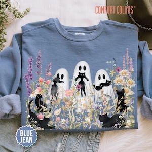 May include: A blue crewneck sweatshirt featuring a graphic print of three white ghosts and several black cats surrounded by colorful wildflowers and lavender. The text COMFORT COLORS is printed in the top right corner and BLUE JEAN in the bottom left.