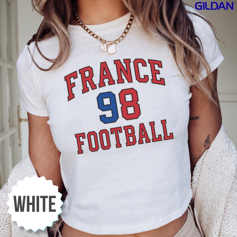 Detailed view of a white cropped baby tee showcasing the retro red and blue France 98 Football logo, paired with a cozy white cardigan. This trendy 90s aesthetic top effortlessly connects past soccer glory with the massive excitement of the upcoming 2026 World Cup. Excellent for casual watch parties with friends at home, cheering at local bars, or sitting in the stadium stands, this premium fan garment is available with reliable US domestic shipping.