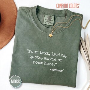 以下が含まれることがあります： 内側のネックラインにComfort Colorsのラベルが付いたモスグリーンのクルーネックTシャツ。前面には「your text, lyrics, quote, movie or poem here.」-optionalという白いテキストがプリントされています。生地の上にはゴールドの星型ペンダントネックレスが置かれています。