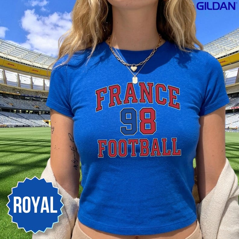 A young woman wears a royal blue Gildan 5000B shirt styled as a 90s Y2K baby tee with necklaces inside an open air stadium. Featuring the classic France 98 Football graphic, this vibrant blue tee pays direct tribute to France's traditional team colors and their historic 1998 triumph. It is the ultimate casual gameday apparel for the 2026 World Cup, ideal for matching with friends at watch parties, family gatherings, or bars, with speedy domestic US shipping.